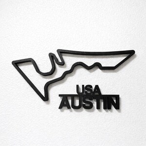 May include: A black wall-mounted sign featuring the outline of the Circuit of the Americas race track in Austin, Texas. Below the track outline, the text USA AUSTIN is displayed in bold, black, capital letters with a textured finish.