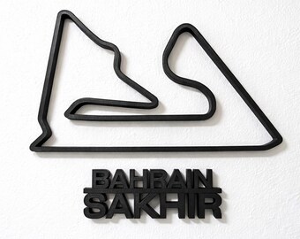 Può includere: Opera d'arte da parete nera raffigurante il circuito di gara del Bahrain e la scritta "BAHRAIN SAKHIR". Il tracciato della pista è una linea continua e il testo è in grassetto senza grazie. L'opera è montata su una parete bianca.