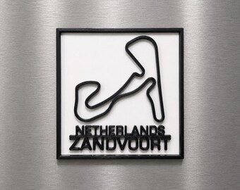 Può includere: Un quadro quadrato da parete con il profilo nero in rilievo del circuito di Zandvoort su sfondo bianco, incorniciato in nero. Il testo NETHERLANDS ZANDVOORT è riportato in lettere nere in rilievo sotto il disegno del tracciato.