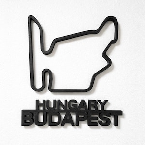 May include: A black wall-mounted sculpture depicting the Hungaroring race track outline. Below the track shape, the words HUNGARY and BUDAPEST are displayed in bold, black, capital letters. The entire piece is constructed from a solid material.