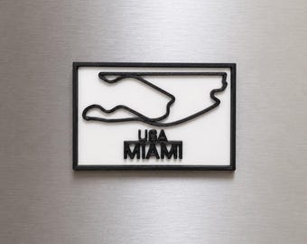 Può includere: Una calamita rettangolare con sfondo bianco e un contorno nero in rilievo del circuito di gara Miami International Autodrome. Sotto il tracciato della pista, il testo USA MIAMI è visualizzato in lettere maiuscole nere in rilievo.
