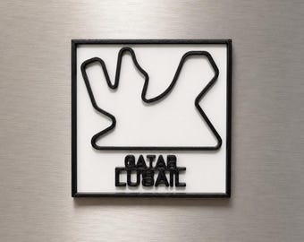 Può includere: Una targa da parete quadrata che presenta un contorno nero in rilievo del circuito di gara Qatar Lusail su sfondo bianco. Il testo QATAR LUSAIL è impresso in lettere nere sotto il tracciato, il tutto racchiuso in una sottile cornice nera.