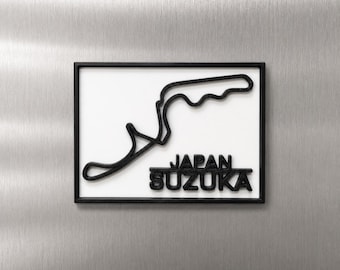 Può includere: Un oggetto da parete rettangolare che presenta il profilo nero del circuito di Suzuka in Giappone su uno sfondo bianco. L'oggetto è racchiuso in una sottile cornice nera e include il testo nero in rilievo JAPAN SUZUKA sotto il tracciato.
