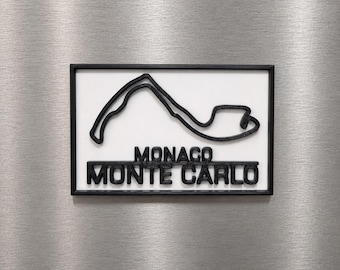 Può includere: Una targa da parete rettangolare con il contorno nero in rilievo del circuito di Monaco su sfondo bianco. Sotto la mappa del circuito, il testo MONACO e MONTE CARLO è visualizzato in lettere maiuscole nere, spesse e in rilievo.