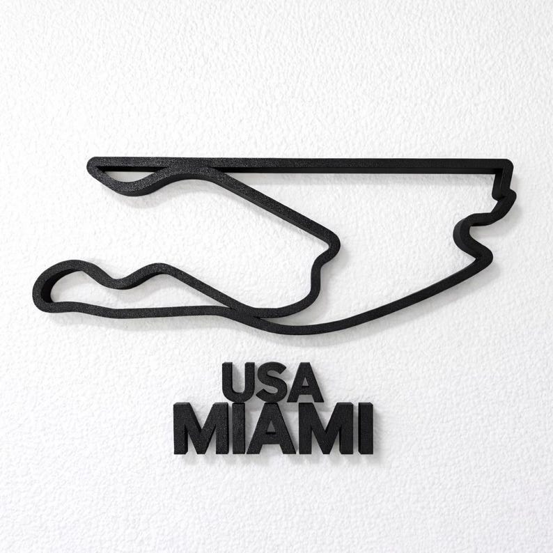 May include: A black, three-dimensional outline of the Miami International Autodrome race track. Below the track layout, the text USA MIAMI is displayed in bold, black, sans-serif capital letters, all set against a textured white background.