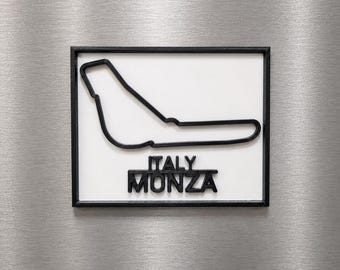 Può includere: Una targa rettangolare da parete che presenta un contorno nero in rilievo del circuito di Monza su sfondo bianco. Sotto il disegno della pista, il testo ITALY MONZA è visualizzato in lettere maiuscole nere, spesse e in rilievo, in una cornice.