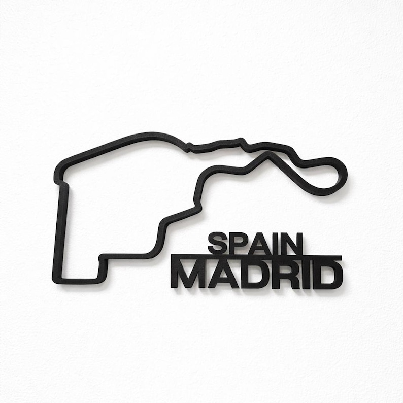 May include: A black wall-mounted sculpture depicting the layout of the Madrid Formula 1 race track. The design features a continuous outline of the circuit with the text SPAIN MADRID positioned in bold, capital letters underneath the track shape.