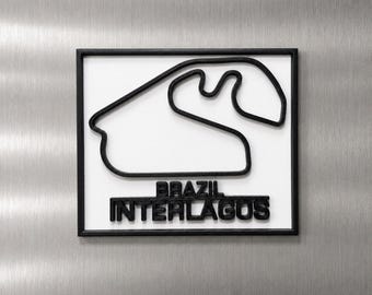 Può includere: Una targa da parete quadrata che presenta un contorno nero in rilievo del circuito di Interlagos in Brasile su sfondo bianco. Il testo BRAZIL INTERLAGOS è visualizzato in lettere nere in rilievo sotto il tracciato, il tutto all'interno di una cornice nera.
