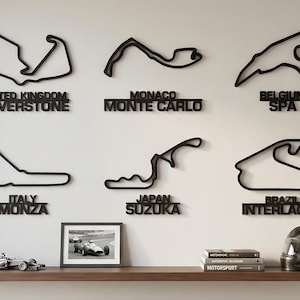 May include: Six black wall-mounted outlines of Formula 1 race tracks including Silverstone, Monte Carlo, Spa, Monza, Suzuka, and Interlagos. Below the tracks, a shelf holds a small framed photograph of a race car, three books, and a helmet.