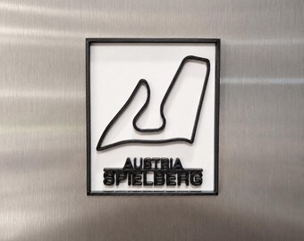 Può includere: Una targa rettangolare da parete con il profilo nero stampato in 3D del circuito di Spielberg, in Austria, su sfondo bianco, incorniciata in nero, con la scritta AUSTRIA SPIELBERG in rilievo nero sotto il tracciato del circuito.