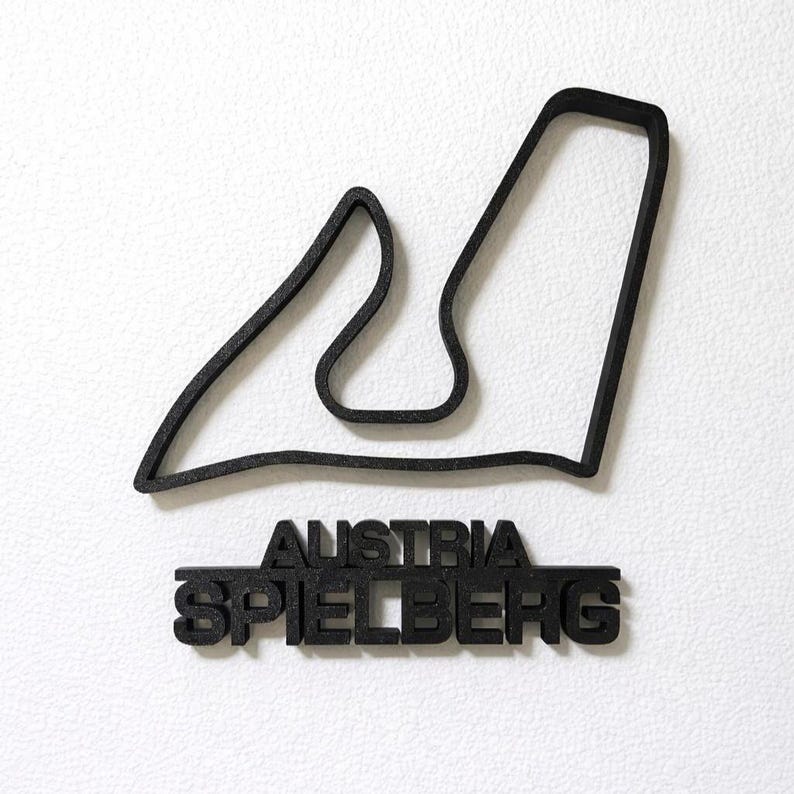 May include: A black 3D-printed wall decoration featuring the outline of the Red Bull Ring racetrack in Spielberg, Austria. Below the track outline, the text AUSTRIA and SPIELBERG are displayed in bold, block capital letters in a matching black finish.