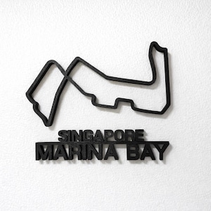 May include: A black, three-dimensional wall-mounted sculpture depicting the Singapore Marina Bay Formula 1 racetrack layout. Below the track outline, the text SINGAPORE MARINA BAY is displayed in bold, black, capital letters.