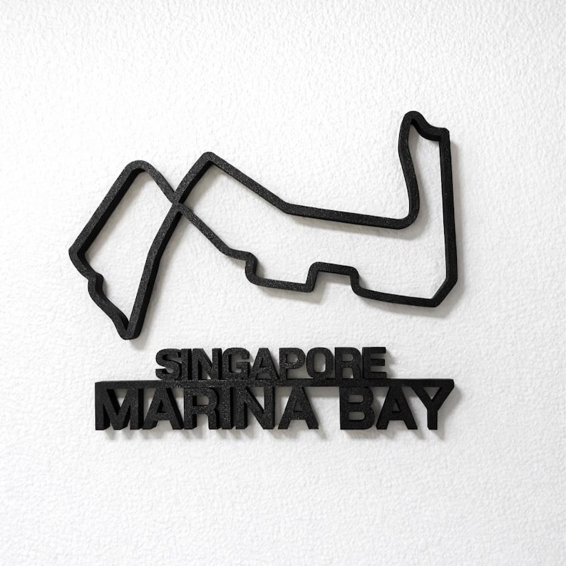 May include: A black, three-dimensional wall-mounted sculpture depicting the Singapore Marina Bay Formula 1 racetrack layout. Below the track outline, the text SINGAPORE MARINA BAY is displayed in bold, black, capital letters.