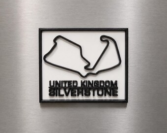 Può includere: Una targa da parete rettangolare con un contorno nero in 3D del circuito di Silverstone su sfondo bianco. Sotto il tracciato, il testo UNITED KINGDOM SILVERSTONE è visualizzato in grassetto nero, il tutto racchiuso in una cornice nera.