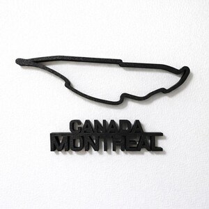 May include: A black, three-dimensional outline of the Circuit Gilles Villeneuve racetrack in Montreal, Canada. Below the track layout, the words CANADA and MONTREAL are displayed in bold, black, block-style capital letters.