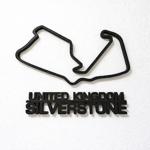 May include: A black, three-dimensional wall decoration featuring the outline of the Silverstone race track in the United Kingdom. Below the track layout, the words UNITED KINGDOM and SILVERSTONE are displayed in bold, black, capital letters.