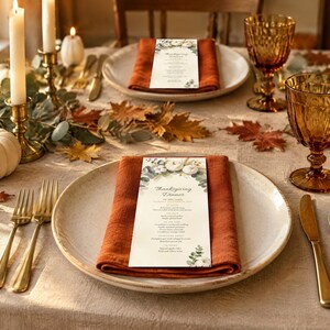 May include: A Thanksgiving dinner menu card with white pumpkins and eucalyptus leaves printed on it, placed on a burnt orange cloth napkin atop a white ceramic plate. The table setting includes gold cutlery, amber glassware, and white candles.