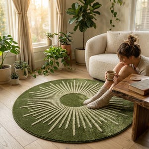May include: A round, olive green rug with a sunburst design in cream. The rug is placed on a wooden floor in a well-lit room with plants and a cream-colored sofa. The rug's diameter is approximately 60 inches.