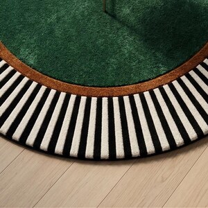 May include: A round rug with a green center, a brown border, and a black and white striped outer edge. The rug is on a light wood floor. The design is modern and geometric.
