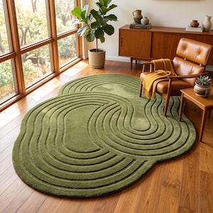 May include: An olive green area rug with an irregular, organic shape and a textured surface featuring carved, concentric, wave-like patterns. The rug is placed on a wooden floor, showcasing a deep, monochromatic tone and a soft, plush pile finish.