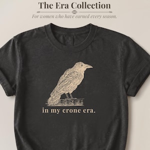 May include: A charcoal gray short-sleeve t-shirt featuring a beige line-art illustration of a crow perched on a branch. Below the bird, the text reads "in my crone era." The shirt has a crew neckline and is tied in a knot at the bottom hem.