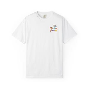 Puede incluir: Camiseta blanca con cuello redondo. La camiseta presenta el texto "Niche, please" en un esquema de colores arcoíris, junto con una cara sonriente y una estrella azul. La camiseta está hecha de un material suave.
