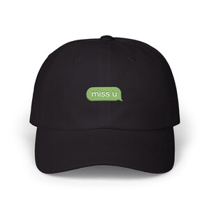 May include: A black baseball cap featuring an embroidered light green speech bubble on the front panel. Inside the speech bubble, the text "miss u" is stitched in white lowercase letters. The cap has a curved brim and a structured crown design.