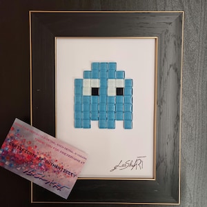 Può includere: Opera d'arte incorniciata con una figura di fantasma blu pixelata, costruita con piccole tessere quadrate. L'opera d'arte è ambientata su uno sfondo bianco all'interno di una cornice nera con un bordo interno dorato. È incluso un certificato di autenticità.