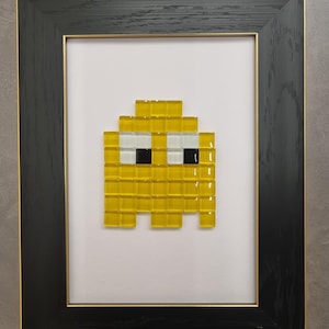 Può includere: Opera d'arte a mosaico incorniciata raffigurante un personaggio fantasma giallo pixelato con occhi bianchi e neri. L'opera è ambientata su uno sfondo bianco all'interno di una cornice nera con un bordo interno dorato. Il mosaico è fatto di piccole tessere quadrate.