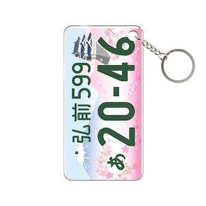 Puede incluir: Un llavero rectangular con un diseño de matrícula japonesa. La placa tiene números verdes y caracteres japoneses sobre un fondo rosa y blanco con flores de cerezo y un paisaje montañoso. Se adjunta una cadena y un anillo plateados.