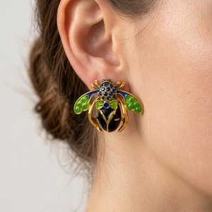 May include: Gold-toned bee earrings with green, blue, and black enamel details. The earrings feature a detailed insect design with a bow tie and wings. The earrings are a unique accessory.