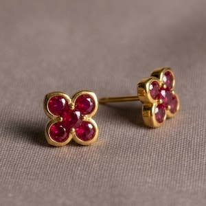 May include: A pair of gold stud earrings, each featuring five round, deep red rubies arranged in a flower-like cluster. The stones are set in polished yellow gold, with one earring shown from the front and the other angled to reveal the post.