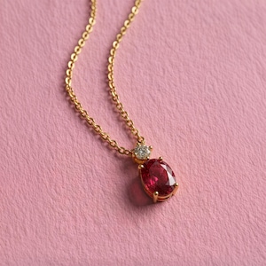May include: A gold chain necklace featuring a pendant with a large oval-cut deep red gemstone and a small round-cut clear stone set directly above it in a gold prong setting, creating a delicate and refined piece of jewelry.