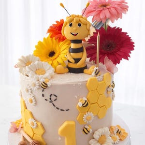 Puede incluir: Una tarta redonda decorada con temática de abejas. Presenta una figura de abeja, detalles de panal, margaritas y el número 1. La parte superior está adornada con gerberas de colores y una vela dorada.