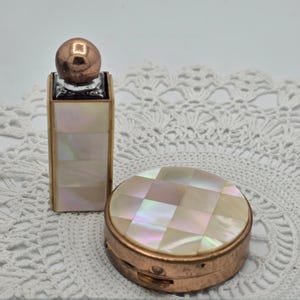 May include: A rectangular perfume bottle and a round compact case, both featuring an iridescent mother-of-pearl inlay. The perfume bottle has a metallic rose gold spherical cap, while the compact case has a matching metallic rose gold base.