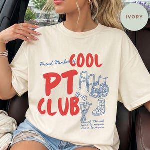 May include: An ivory short-sleeved t-shirt featuring the red text "Proud Member of COOL PT CLUB" and blue line art illustrations of a walker, a wheelchair, a dumbbell, crutches, and a medical boot, with the text "Physical Therapist fueled by purpose, driven by progress."