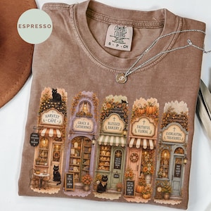 May include: A brown t-shirt featuring a graphic print of five storefronts labeled Harvest Cafe, Grace & Gatherings, Blessed Bakery, Faithful Floral, and Everlasting Treasures, with black cats and autumn decorations, and a silver chain necklace.
