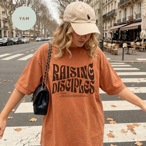 May include: A beige baseball cap with a small dark logo and an oversized burnt orange t-shirt featuring the dark brown text RAISING DISCIPLES in a wavy font, along with smaller text below, paired with a black quilted shoulder bag with a chain strap.