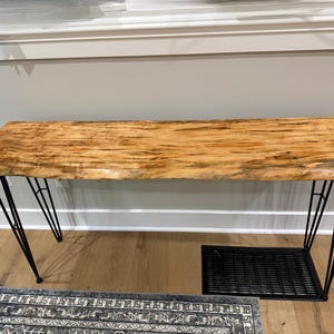 May include: A rectangular wooden bench with a natural wood grain finish. The bench has black metal hairpin legs and a black metal shelf underneath. The wood has a light brown color with darker streaks.