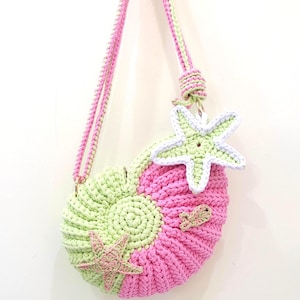Könnte beinhalten: Handgefertigte Häkeltasche im Muscheldesign mit rosa und hellgrünen Abschnitten. Die Tasche hat einen rosa und hellgrünen Riemen und ist mit Seesternen und einem kleinen Fisch verziert. Die Tasche hängt an einem Haken.