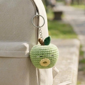 May include: A beige canvas backpack with a light green crocheted apple keychain attached to the side zipper. The apple features a small brown stem, a single dark green leaf, and a circular beige mouth detail stitched onto the front surface.