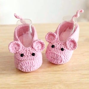 May include: A pair of light pink crocheted baby booties designed to resemble pigs. Each bootie features two round ears, a snout-like front section, and a small curly tail at the back. The eyes are made of small, round, black beads attached to the front.