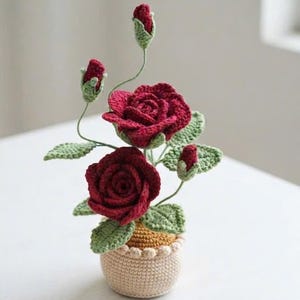 Könnte beinhalten: Eine gehäkelte Rosenpflanze mit zwei voll erblühten dunkelroten Rosen und drei kleineren Rosenknospen an dünnen grünen Stielen. Die Pflanze hat strukturierte grüne Blätter und steht in einem kleinen, runden, beigefarbenen gehäkelten Topf.