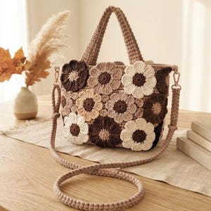 May include: A crochet handbag featuring a floral pattern made of individual circular motifs in shades of cream, beige, and dark brown. The bag has two short top handles and a long, detachable shoulder strap, all crafted from matching yarn.