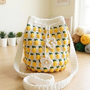 May include: A white crochet bucket bag featuring a repeating pattern of yellow lemon-shaped bobbles with green leaves. The bag has a drawstring closure with two white crocheted flower accents and a long, wide white crocheted shoulder strap.