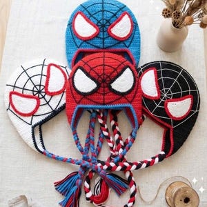 Puede incluir: Tres gorros de ganchillo con patr&oacute;n de telara&ntilde;a y grandes ojos blancos con bordes rojos. Los gorros son azules, rojos y negros, cada uno con largas orejeras trenzadas que terminan en borlas coloridas. El dise&ntilde;o imita una m&aacute;scara.