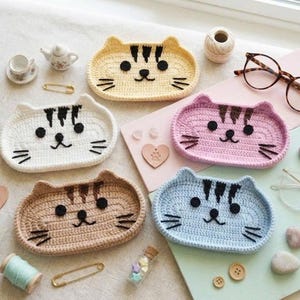 May include: Five oval crochet trays shaped like cat faces in yellow, white, tan, pink, and light blue. Each tray features black embroidered eyes, noses, whiskers, and forehead stripes. The trays are arranged with small craft items like spools of thread, buttons, safety pins, and heart-shaped charms.