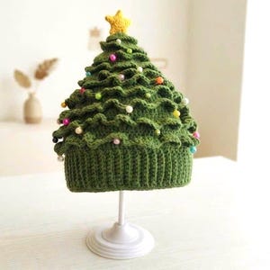 Può includere: Un berretto all'uncinetto verde a forma di albero di Natale con bordo a coste. Il berretto presenta strati a balze decorati con piccole perline multicolori ed è sormontato da una stella all'uncinetto gialla. Il berretto poggia su un supporto bianco.