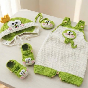 May include: A crochet set featuring a green and white hat with a yellow crown, two green shoes, a small green circular pouch, and a white and green romper. Each piece displays a cartoon frog face with large white eyes, black pupils, and pink cheeks.