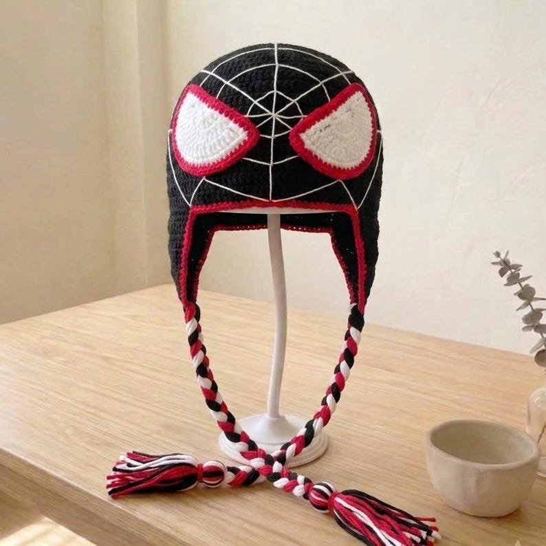 Puede incluir: Un gorro de ganchillo negro con detalles de telara&ntilde;a blancos y dos grandes parches oculares blancos delineados en rojo. El gorro cuenta con orejeras con largas borlas trenzadas de hilo negro, blanco y rojo que cuelgan de cada lado.
